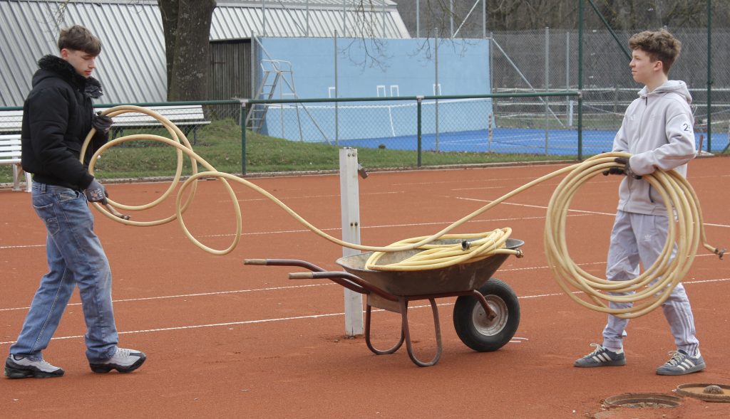 Das Bild zeigt zwei Jugendliche, die einen Wasserschlauch für die Tennisplätze abladen